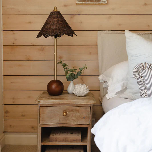 Wooden nightstand with lamp and decor items next to a bed with pillows against a wooden wall.