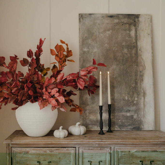 Decorative setup with a vase of red flowers, candles, and pumpkins on a wooden surface.