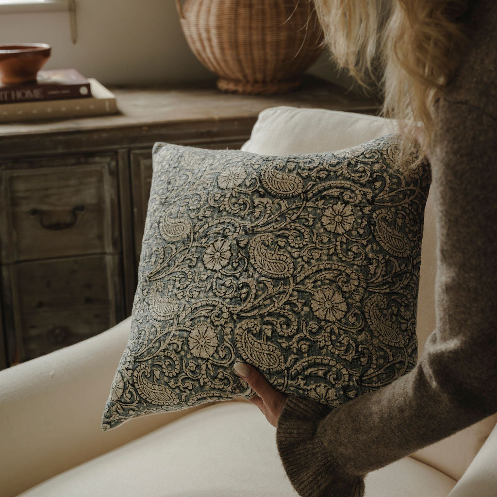 Person holding a patterned pillow in a cozy room with wooden furniture and a vase.