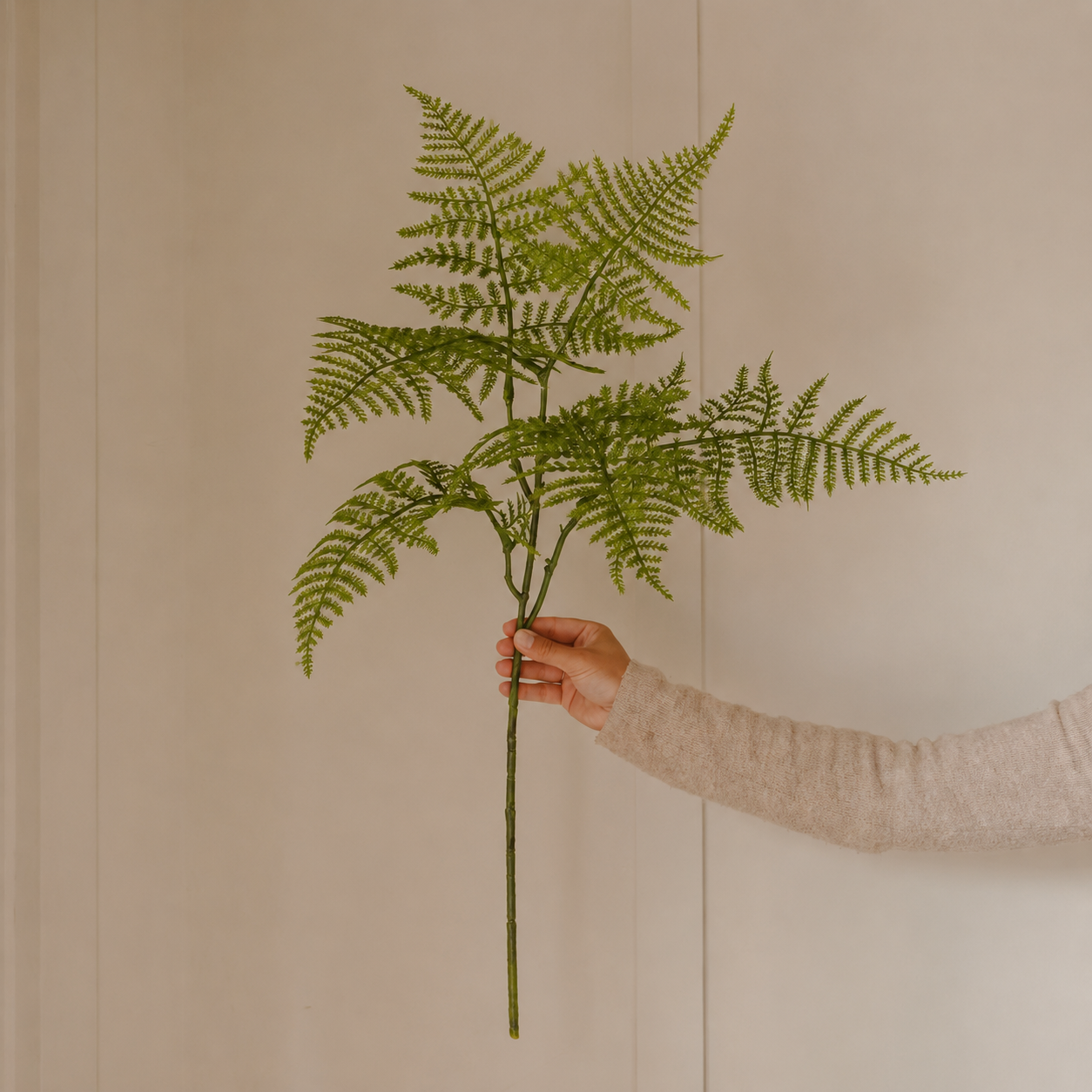Artificial fern stem held by hand against a soft neutral beige panelled background
Faux green fern branch styled in hand with warm beige wall background
Decorative fern stem with delicate green fronds held in front of a neutral indoor backdrop
Single artificial fern spray with fine leafy fronds displayed against a soft cream panelled wall
Botanical faux fern stem held in a hand, styled on a warm neutral background