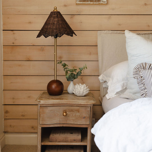 Wooden nightstand with lamp and decor items next to a bed with pillows against a wooden wall.