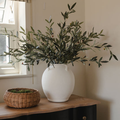 White vase with greenery on a wooden surface next to a window