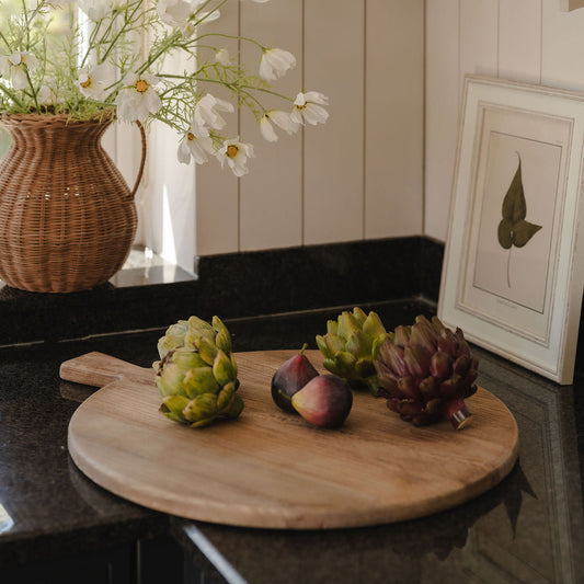 Wooden cutting board with fruits and a vase of flowers on a kitchen counter.